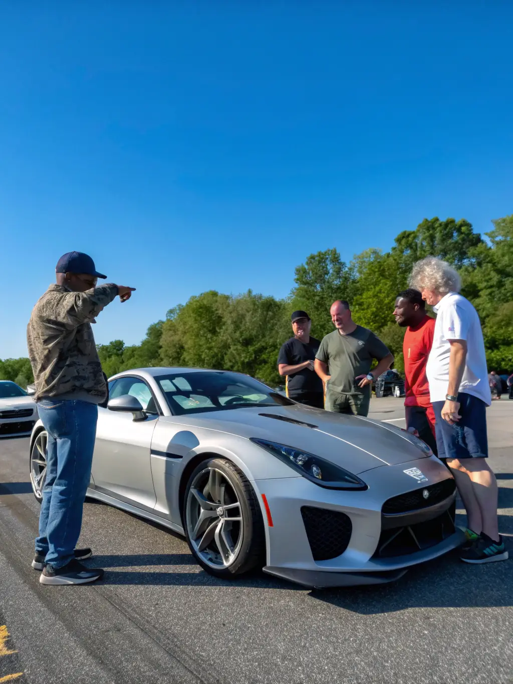 A diverse group of people, including older and younger generations, gathered around a vintage car, discussing its history and mechanics.